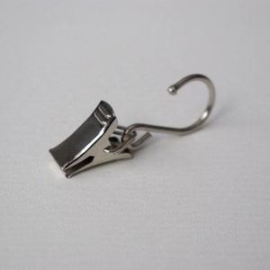 May include: A silver metal curtain clip with a hook. The clip has a spring-loaded mechanism to hold the curtain in place.
