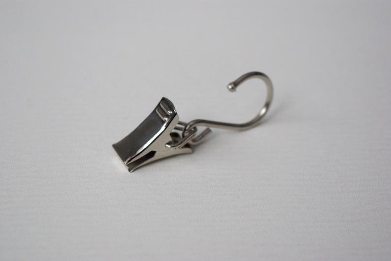 May include: A silver metal curtain clip with a hook. The clip has a spring-loaded mechanism to hold the curtain in place.