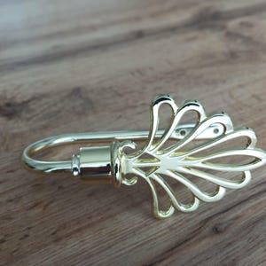 May include: A gold-colored curtain tieback with a decorative fan-shaped design. The tieback has a curved hook and a detailed, ornate fan-like embellishment. The metal appears to be polished, reflecting light. The background is a wooden surface.