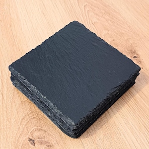 May include: A stack of four square, black slate coasters with rough, textured edges. The coasters are arranged on a light brown wooden surface, creating a contrast in colour and texture. The slate appears to be natural stone.
