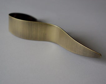 May include: A brass-colored, curved metal object with a brushed finish. The design features a wide, rounded top that tapers to a pointed end. The object is set against a plain, light background, highlighting its form and texture.