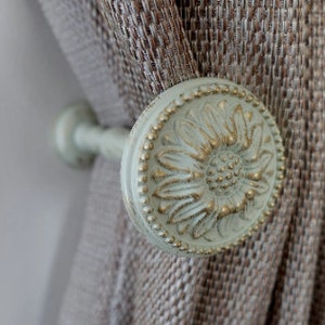 May include: A white and gold decorative curtain tieback with a floral design. The tieback is attached to a brown curtain.
