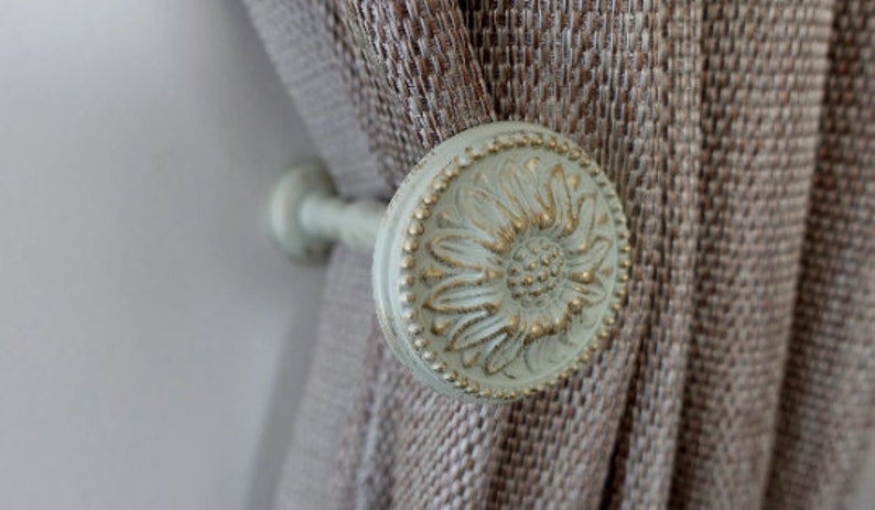 May include: A white and gold decorative curtain tieback with a floral design. The tieback is attached to a brown curtain.