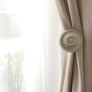 May include: A white, decorative curtain tieback with a floral design. The tieback is attached to a beige curtain.
