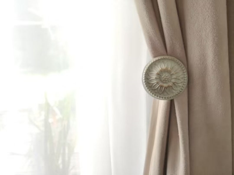 May include: A white, decorative curtain tieback with a floral design. The tieback is attached to a beige curtain.