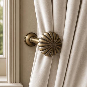 May include: A brass curtain holdback featuring a round, fluted, shell-like design with a central mounting post. The metal hardware is finished in an antique bronze tone and secures a heavy, light beige velvet curtain panel against a wall.
