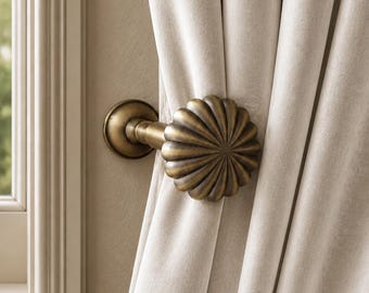 May include: A brass curtain holdback featuring a round, fluted, shell-like design with a central mounting post. The metal hardware is finished in an antique bronze tone and secures a heavy, light beige velvet curtain panel against a wall.