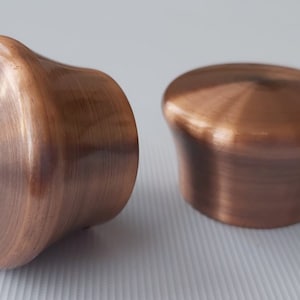 May include: Two copper-coloured cabinet knobs with a brushed finish. The knobs have a rounded top and a curved, flared base. The metal has a warm, reddish-brown hue, and the surface shows circular brush marks.