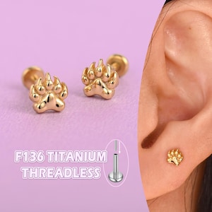 Puede incluir: Pendientes sin rosca de titanio color oro con forma de huella de pata. Los pendientes se muestran sobre un fondo morado claro, con el texto "F136 TITANIUM THREADLESS" en blanco. Un pendiente se muestra en una oreja.