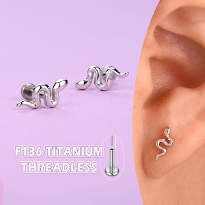 May include: Silver-colored snake-shaped ear piercing studs. The image shows two studs and one stud in an ear. The text "F136 TITANIUM THREADLESS" is also visible.