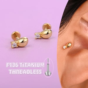 May include: Gold-colored, threadless titanium earrings with a teardrop design and a small, clear gemstone accent. The earrings are shown on a purple background, with the text "F136 TITANIUM THREADLESS" in a white font.