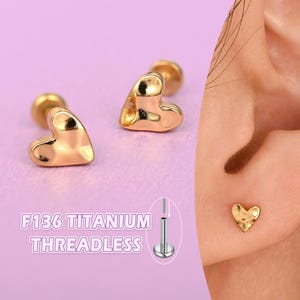 May include: Two gold-colored, heart-shaped earrings with a textured surface. The earrings are threadless and have a post. The image also shows the text "F136 TITANIUM THREADLESS".