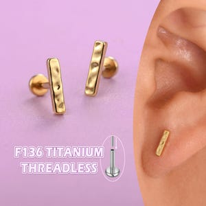 May include: Gold-colored, threadless titanium earrings with a hammered bar design. The earrings are shown on a purple background, with the text "F136 TITANIUM THREADLESS" in the lower left corner. One earring is shown in an ear piercing.