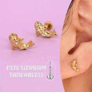 May include: Gold-coloured fish-shaped stud earrings. The earrings are shown on a light purple background, with a close-up of the earring in a pierced ear. The text "F136 TITANIUM THREADLESS" is also shown.
