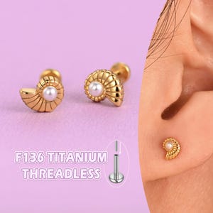 May include: Gold-colored, threadless titanium earrings in a seashell design, each with a small, white pearl. The earrings are shown on a purple background, with the text "F136 TITANIUM THREADLESS".
