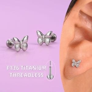 May include: Silver-colored butterfly-shaped threadless earrings. The earrings are shown in a close-up shot, with the text "F136 TITANIUM THREADLESS" in the image. The butterfly design is detailed and delicate, with visible wing patterns. The earrings are shown on a person's ear.