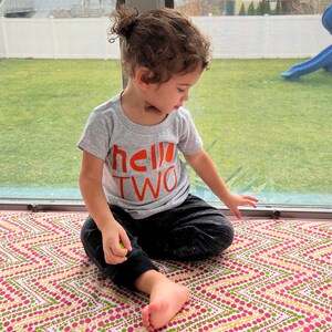 May include: A toddler wearing a grey t-shirt with the words "hello two" in orange lettering. The child is sitting on a colourful patterned rug.