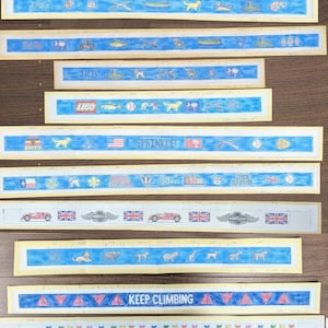 May include: A collection of ten horizontal paper strips featuring hand-drawn illustrations on blue backgrounds. The designs include various icons, animals, vehicles, flags, sports equipment, and text such as "SPRINKLE", "KEEP CLIMBING", and "CDK".