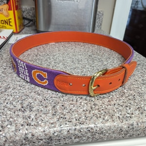 May include: A purple and orange woven belt with a gold buckle. The belt has the years 1981, 2016, and 2018 embroidered on it, along with a large letter "C".