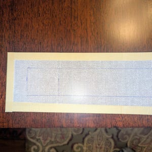 May include: A rectangular needlepoint canvas with a grid pattern, featuring a light yellow border. The canvas has a drawn rectangular outline in the center. The canvas is placed on a dark brown wooden surface.