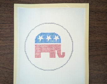 May include: A white canvas with a red, white, and blue elephant design in a circle. The elephant is facing left and has a star-studded blue background. The canvas is framed with a yellow border.