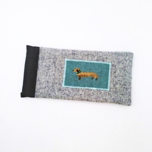 May include: A rectangular, grey felt case with a black accent on one end. A teal rectangle with a dachshund illustration is sewn onto the case. The dachshund is brown with a green collar. The case is likely for eyeglasses or a phone.