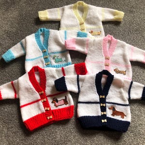 Puede incluir: Cinco cárdigans de bebé hechos a mano en varios colores, incluyendo blanco con ribete rojo, blanco con ribete azul y blanco con ribete rosa. Cada cárdigan presenta un aplique de perro salchicha y botones de madera. Los cárdigans son de punto.