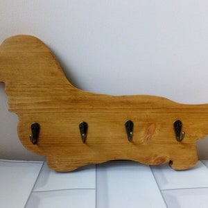 May include: A wooden key holder in the shape of a dachshund dog, featuring four bronze hooks. The key holder is crafted from light-coloured wood and is mounted on a white and grey tiled surface.