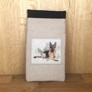 May include: A rectangular, beige fabric case with a black top. A rectangular patch on the front features a German Shepherd dog illustration. The dog is lying down, with black and tan fur, on a light green background.