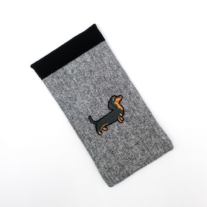 May include: A gray eyeglasses case with a black top and a dachshund dog patch. The case is rectangular and made of a textured fabric. The dachshund is black and brown with an orange outline.