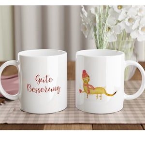 Könnte beinhalten: Zwei weiße Keramikbecher mit rotem und weißem Design. Ein Becher hat den Text "Gute Besserung" darauf. Der andere Becher hat eine Comic-Illustration eines gelben Tieres mit roter Mütze und Schal.