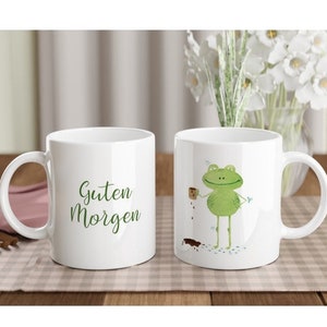 Puede incluir: Dos tazas de cerámica blanca, una con una ilustración de rana verde y la otra con el texto "Guten Morgen".