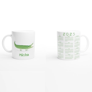 Puede incluir: Taza de cerámica blanca con una ilustración de un cocodrilo verde y el nombre "Micha" impreso en la parte delantera. La parte posterior de la taza presenta un calendario verde 2025.