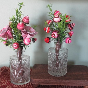May include: Two decorative floral arrangements in clear glass vases. Each arrangement features green stems with pink and red spiral-shaped ornaments and paper flowers. The vases have a diamond pattern and sit on a wooden surface.