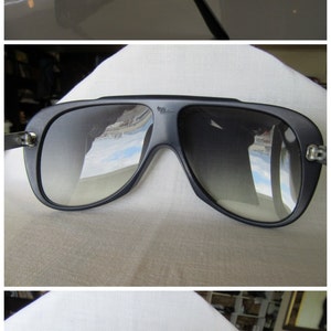 K&ouml;nnte beinhalten: Schwarze Aviator-Sonnenbrille mit silbernem Rahmen und grauen Gl&auml;sern. Die Sonnenbrille spiegelt sich auf einer wei&szlig;en Oberfl&auml;che.