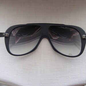 K&ouml;nnte beinhalten: Schwarze Aviator-Sonnenbrille mit dunkelgrauem Verlaufsglas. Die Sonnenbrille hat einen dicken schwarzen Rahmen und eine Metallbr&uuml;cke.