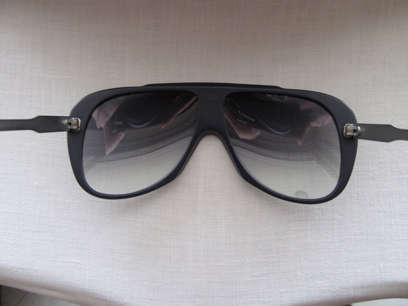 K&ouml;nnte beinhalten: Schwarze Aviator-Sonnenbrille mit dunkelgrauem Verlaufsglas. Die Sonnenbrille hat einen dicken schwarzen Rahmen und eine Metallbr&uuml;cke.