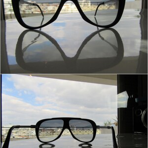 K&ouml;nnte beinhalten: Eine schwarze Aviator-Sonnenbrille mit reflektierendem Glas. Die Sonnenbrille spiegelt einen bew&ouml;lkten Himmel und Geb&auml;ude im Hintergrund wider.