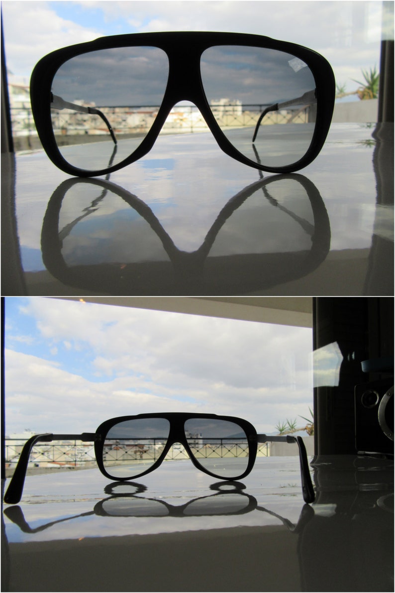 K&ouml;nnte beinhalten: Eine schwarze Aviator-Sonnenbrille mit reflektierendem Glas. Die Sonnenbrille spiegelt einen bew&ouml;lkten Himmel und Geb&auml;ude im Hintergrund wider.