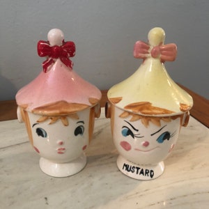 May include: A set of two vintage ceramic salt and pepper shakers. The shakers are shaped like children's heads with painted faces and hats. The girl shaker has a pink hat with a red bow and the boy shaker has a yellow hat with a pink bow. The boy shaker has the word "MUSTARD" written on the bottom.