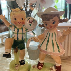 May include: Two vintage ceramic figurines of children. The boy is wearing a green and white striped shirt and green shorts. He is holding a blue and white ball. The girl is wearing a white dress with green and white stripes. She is holding a blue bucket. Both figurines are standing on green bases.