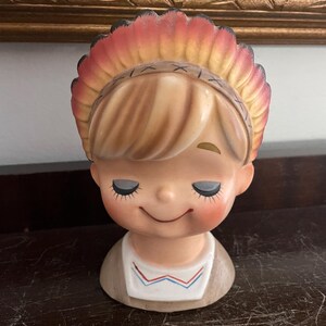 May include: A ceramic figurine of a smiling child with closed eyes, wearing a red and yellow headdress. The figurine is a vintage style and has a brown and white painted design.