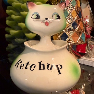 May include: A white ceramic condiment jar shaped like a cat with green accents on the ears and base. The jar features the word Ketchup printed in black script lettering on the front. The cat head serves as a lid for the rounded container.
