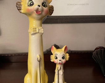May include: Two ceramic cat figurines with large eyes and a yellow and white color scheme. The larger cat is standing on its hind legs with a gold chain around its neck. The smaller cat is sitting with a gold chain around its neck.