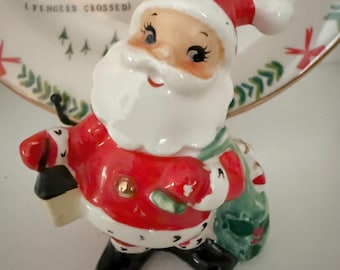 May include: A ceramic Santa Claus figurine with a red suit, black boots, and a white beard. He holds a lantern and a green sack. The background includes a plate with the words "SNOW DAY (FINGERS CROSSED)" and winter-themed illustrations.