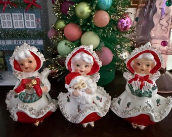 Könnte beinhalten: Drei Vintage-Weihnachtsfiguren von Frauen in roten und weißen Kleidern und Hauben. Jede Figur hält einen festlichen Gegenstand, wie einen Kranz oder ein Geschenk. Im Hintergrund steht ein kleiner Weihnachtsbaum mit bunten Ornamenten.