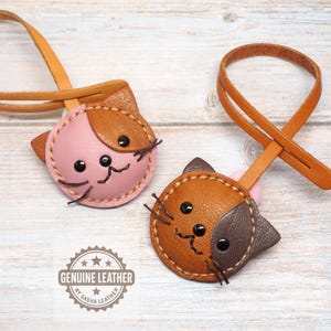 May include: Two leather cat-shaped keychains. One is pink and brown, the other is brown and dark brown. Both have black eyes and stitched details. Each keychain has a loop for attaching to keys. The words "GENUINE LEATHER" are printed on the image.