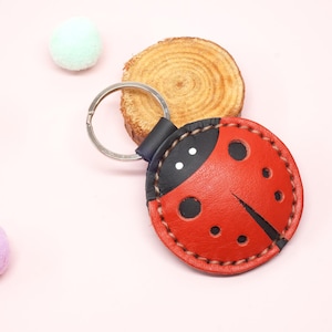 Puede incluir: Un llavero de cuero rojo y negro con forma de mariquita con un llavero plateado. La mariquita tiene lunares negros y una cabeza negra. El llavero está sobre un fondo rosa con tres pompones pastel y una rodaja de madera.