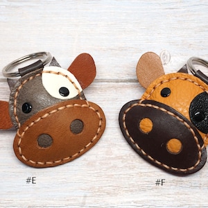 May include: Two cow-shaped keychains with metal rings. Keychain #E is grey and brown with a white patch and black eyes. Keychain #F is yellow and dark brown with a black patch and black eyes. Both feature visible stitching and are labeled #E and #F.