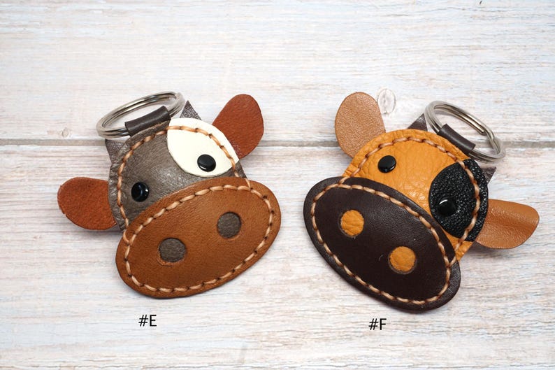 May include: Two cow-shaped keychains with metal rings. Keychain #E is grey and brown with a white patch and black eyes. Keychain #F is yellow and dark brown with a black patch and black eyes. Both feature visible stitching and are labeled #E and #F.